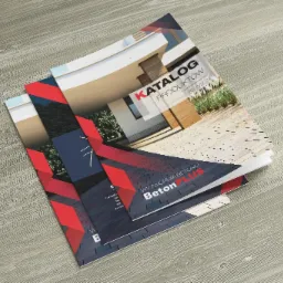 Stos katalogów produktowych firmy Beton Plus na drewnianym tle, prezentujących kostkę brukową i architekturę.