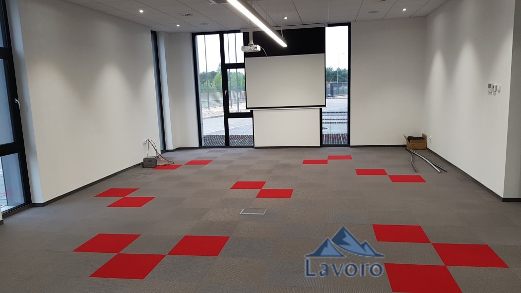 Wnętrze biurowe z szarą wykładziną w czerwone kwadraty, widoczny projektor i ekran, częściowo zamontowana listwa przypodłogowa przy ścianie.