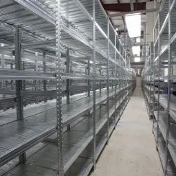 Puste, metalowe regały magazynowe z perforowanymi słupkami, widok w głąb korytarza, oświetlenie sufitowe.