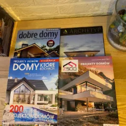 Katalogi i magazyny z projektami domów ułożone na drewnianej półce, widoczne okładki z wizualizacjami nowoczesnych domów jednorodzinnych i basenów.