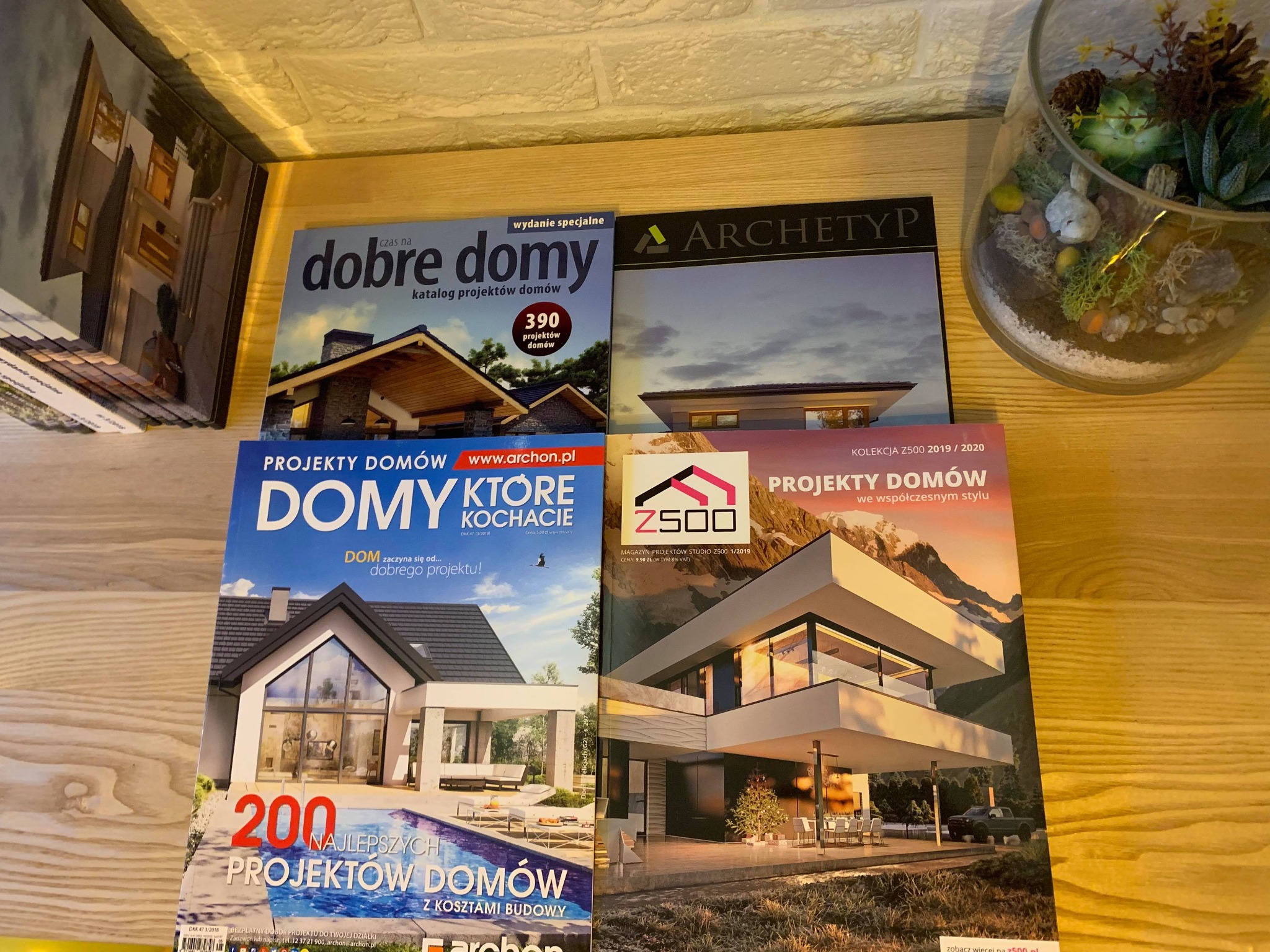 Katalogi i magazyny z projektami domów ułożone na drewnianej półce, widoczne okładki z wizualizacjami nowoczesnych domów jednorodzinnych i basenów.