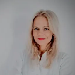 Portret uśmiechniętej blondynki w białym fartuchu medycznym na jasnym tle.