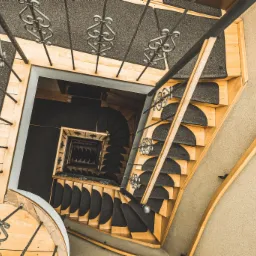 Widok z góry na drewniane schody spiralne z czarnymi nakładkami dywanowymi i metalową balustradą z ozdobnymi elementami.