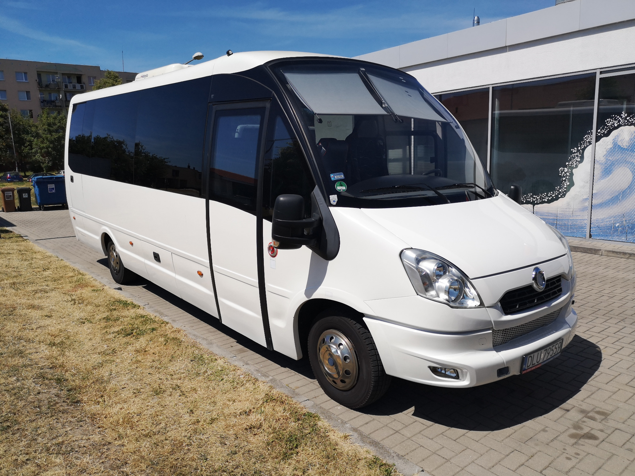 Biały minibus z przyciemnianymi szybami, zaparkowany na chodniku z kostki brukowej, z widocznym budynkiem w tle.