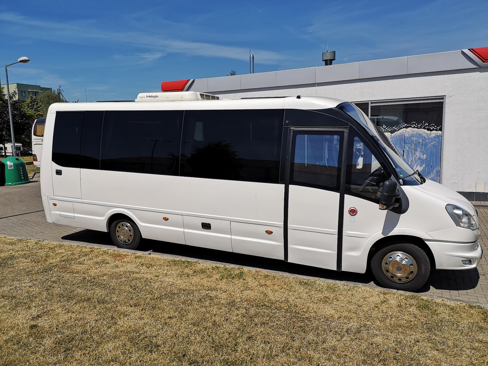 Biały minibus z ciemnymi szybami zaparkowany na trawie, widoczny klimatyzator Webasto na dachu i budynek z czerwonym dachem w tle.
