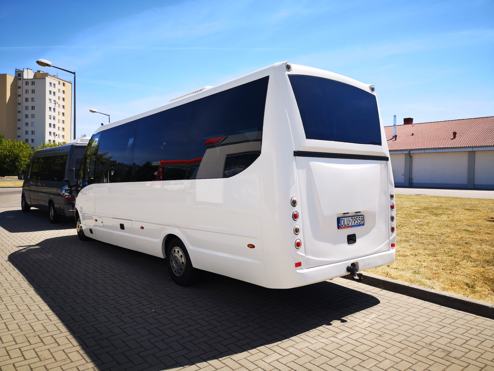 Biały minibus z ciemnymi szybami, zaparkowany na kostce brukowej w słoneczny dzień; w tle budynek mieszkalny i trawnik.