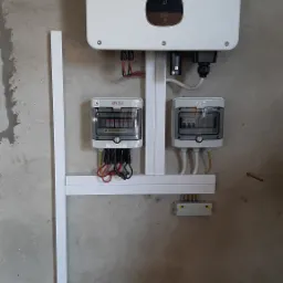 Biały falownik Growatt zamontowany na szarej ścianie, z dwoma rozdzielnicami elektrycznymi ETI podłączonymi przewodami w kolorach czerwonym, czarnym i żółto-zielonym, przymocowany do ściany za...