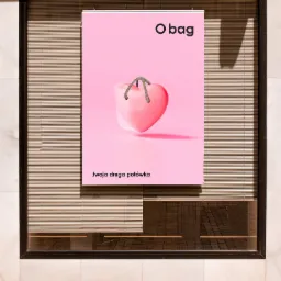 Różowy plakat reklamowy O bag z motywem serca i hasłem '.twoja druga połówka' zawieszony na ścianie z żaluzjami.