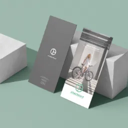 Dwa pionowe foldery reklamowe na tle miętowej powierzchni i białych bloków. Jeden folder jest szary z logo i adresem strony internetowej, drugi przedstawia kobietę na rowerze elektrycznym i logo...