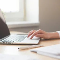 Kobieta w białej koszuli pracuje na laptopie przy biurku z notatnikiem i piórem, kubek kawy w tle.