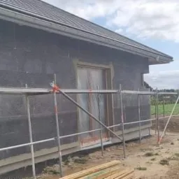 Budowa domu jednorodzinnego z czarnym styropianem na ścianach, widoczne rusztowanie i przygotowane deski, w tle pole i drzewa.