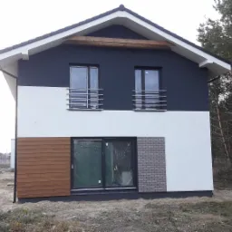 Dwukondygnacyjny dom z płaskim dachem, z fragmentami elewacji wykończonymi drewnem i cegłą, widoczne okna z barierkami, na tle lasu i budowy.