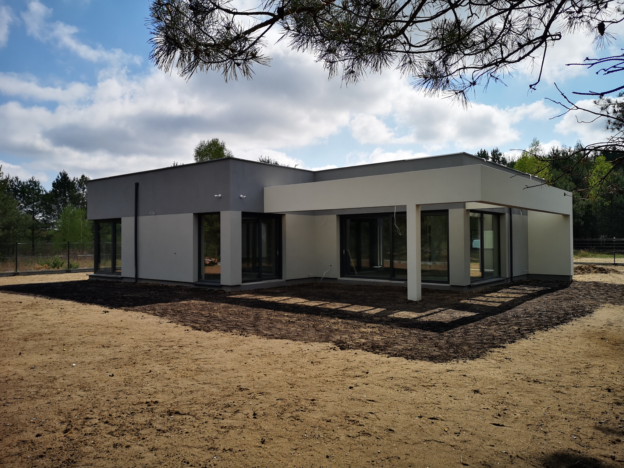 Nowoczesny parterowy dom z płaskim dachem, wykończony w odcieniach szarości i bieli, otoczony przygotowanym gruntem pod trawnik, widok z zewnątrz w słoneczny dzień.