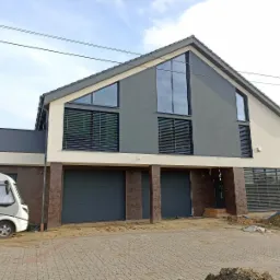 Dwukondygnacyjny dom z szarej i kremowej elewacji z dużymi oknami i dwoma garażami, widoczny kamper na podjeździe z kostki brukowej.