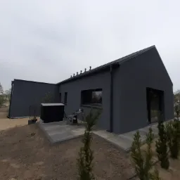 Nowoczesny, parterowy dom w ciemnej kolorystyce z płaskim dachem, otoczony młodymi drzewkami iglastymi, z widocznym tarasem i meblami ogrodowymi.