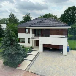 Nowoczesny dwukondygnacyjny dom z ciemną dachówką, białymi ścianami i elementami z ciemnego drewna, widok z góry na podjazd z kostki brukowej i otwarty garaż z zaparkowanym samochodem.