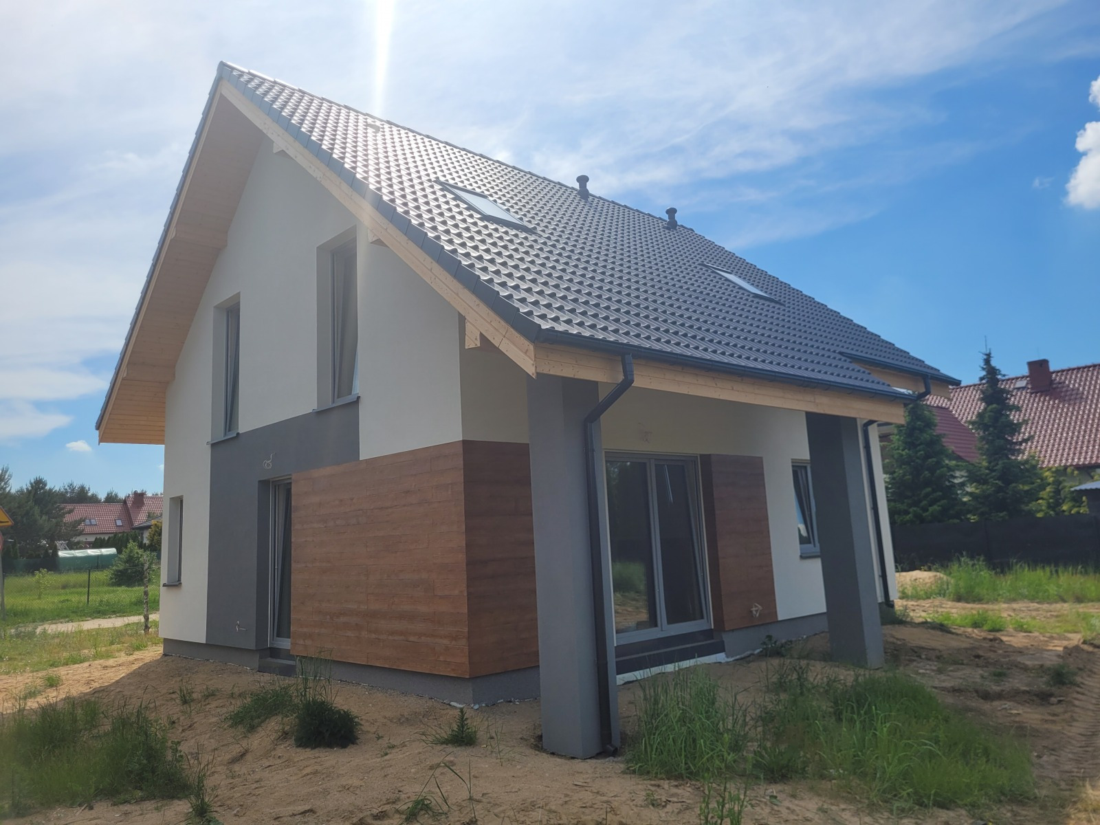 Nowoczesny dom jednorodzinny w Krakowie z ciemną dachówką, jasną elewacją i drewnianymi panelami, widziany w słoneczny dzień.