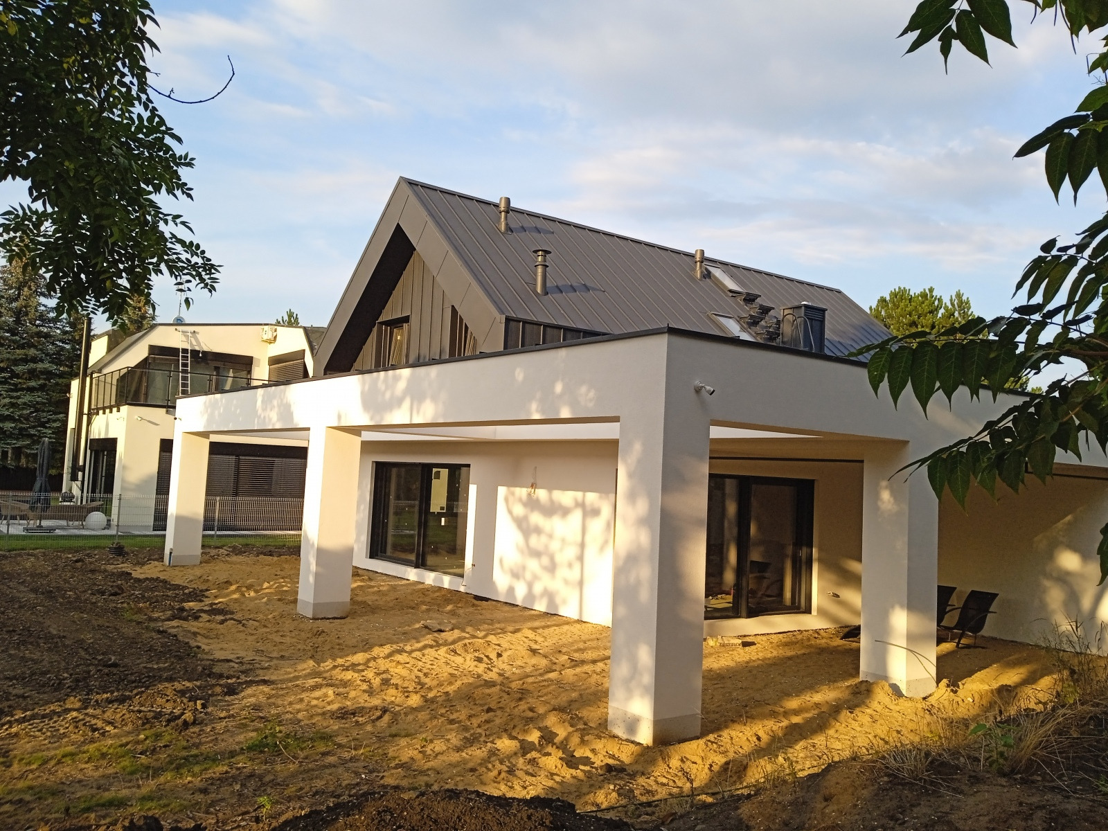 Nowoczesny dom murowany z płaskim dachem i zadaszonym tarasem w trakcie wykańczania ogrodu, widoczne świeżo nawieziona ziemia i piasek.