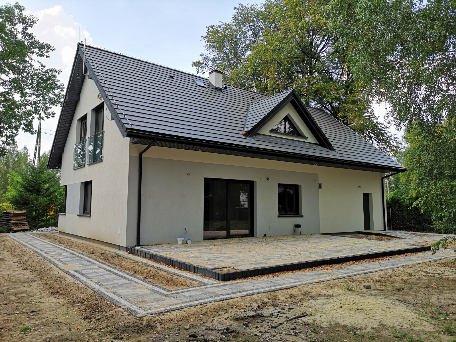 Nowoczesny dom jednorodzinny z szarą dachówką i elewacją w odcieniach bieli i szarości, widoczny taras wyłożony kostką brukową w trakcie prac wykończeniowych, ogród w tle.