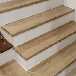 Wykończone schody z płytek imitujących drewno, białe podstopnice, widoczne fugi i fragment podłogi z ciemnych płytek.