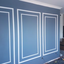 Wamas usługi remontowe budowlane - Ściana w kolorze gołębiej szarości z dekoracyjnymi białymi listwami tworzącymi panele, widoczne gniazdka elektryczne i narzędzia na podłodze.