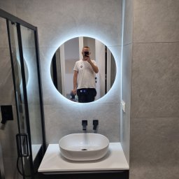 Wamas usługi remontowe budowlane - Nowoczesna łazienka z szarą płytką imitującą kamień, okrągłe lustro z podświetleniem LED i białą umywalką nablatową z czarną armaturą. Kabina prysznicowa z czarnymi profilami.
