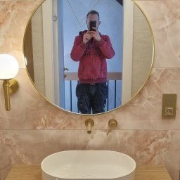 Wamas usługi remontowe budowlane - Ukończona łazienka z umywalką nablatową, złotą armaturą i lustrem. Widoczne płytki imitujące marmur i pracownik robiący zdjęcie w lustrze.