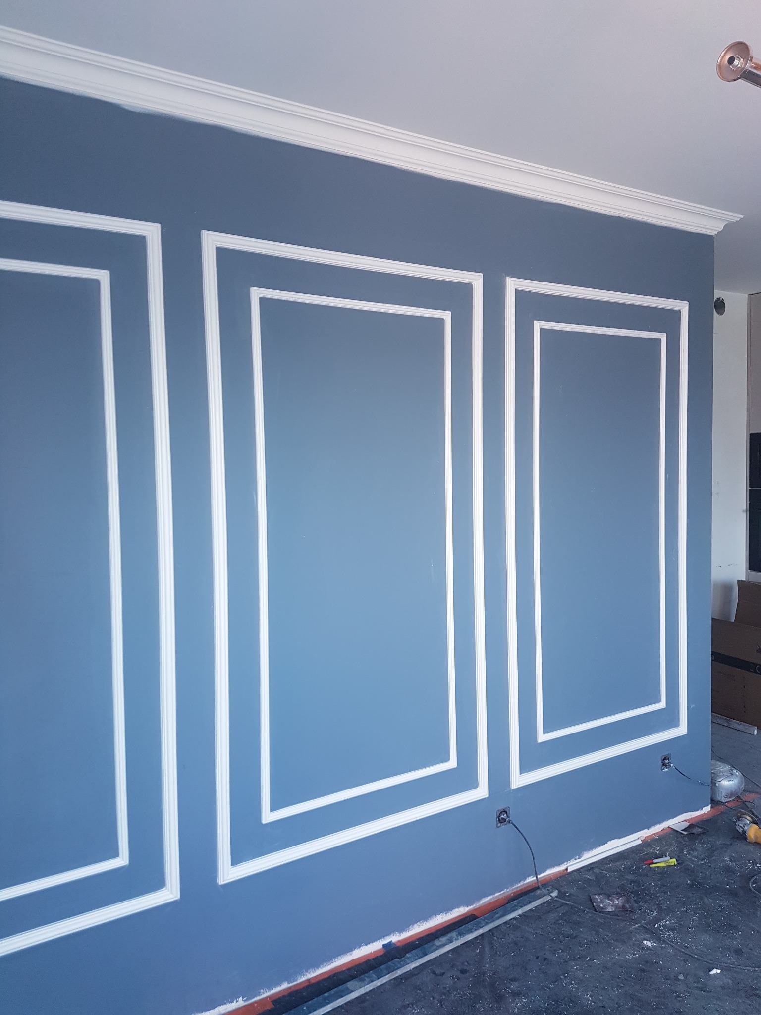 Ściana w kolorze gołębiej szarości z dekoracyjnymi białymi listwami tworzącymi panele, widoczne gniazdka elektryczne i narzędzia na podłodze.