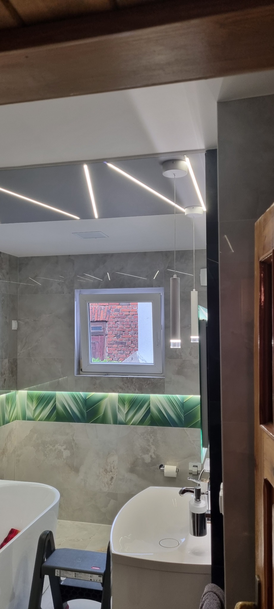 Nowoczesna łazienka z szarymi płytkami imitującymi marmur, podświetlanym sufitem z geometrycznym wzorem, zielonym pasem dekoracyjnym z motywem liści pod oknem oraz wiszącymi lampami cylindrycznymi,...