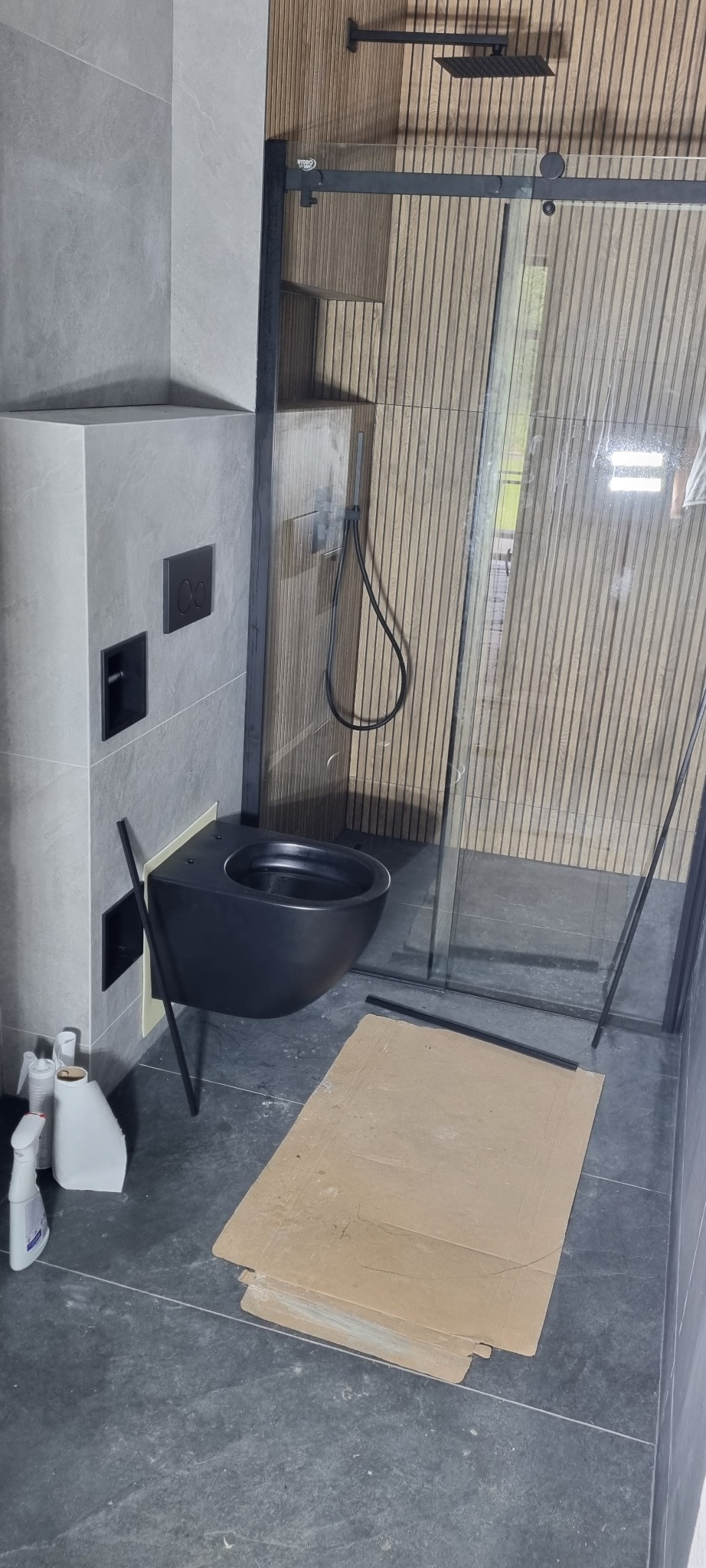Nowoczesna łazienka w trakcie wykańczania: czarna podwieszana toaleta, szklana kabina prysznicowa z czarnymi ramami, drewniana boazeria na ścianie, szare płytki podłogowe i zabezpieczenia z kartonu...