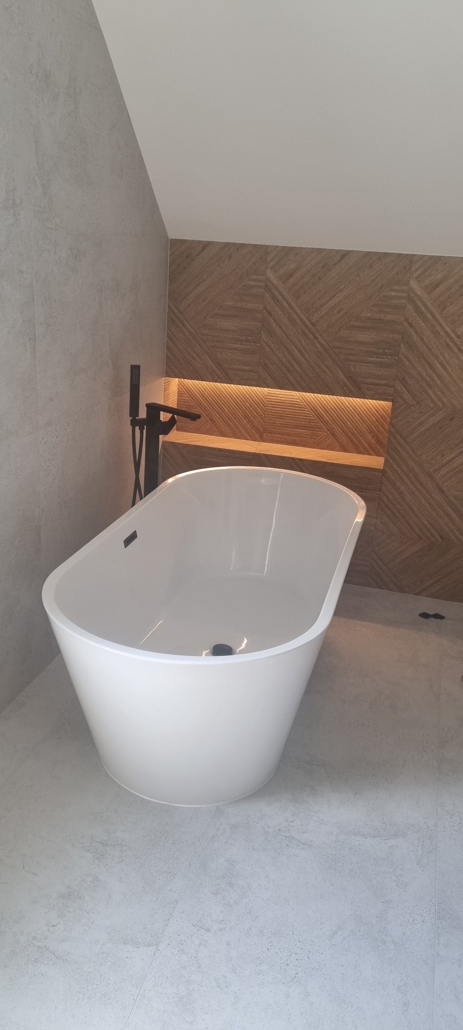 Wolnostojąca, biała wanna z czarną armaturą w nowoczesnej łazience z drewnianą ścianą podświetloną paskiem LED.
