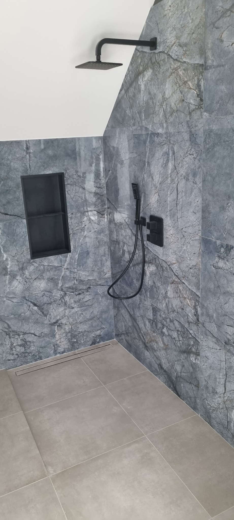 Wykończona kabina prysznicowa z szarymi płytkami imitującymi marmur, czarną armaturą i odpływem liniowym w podłodze z jasnych, kwadratowych płytek.