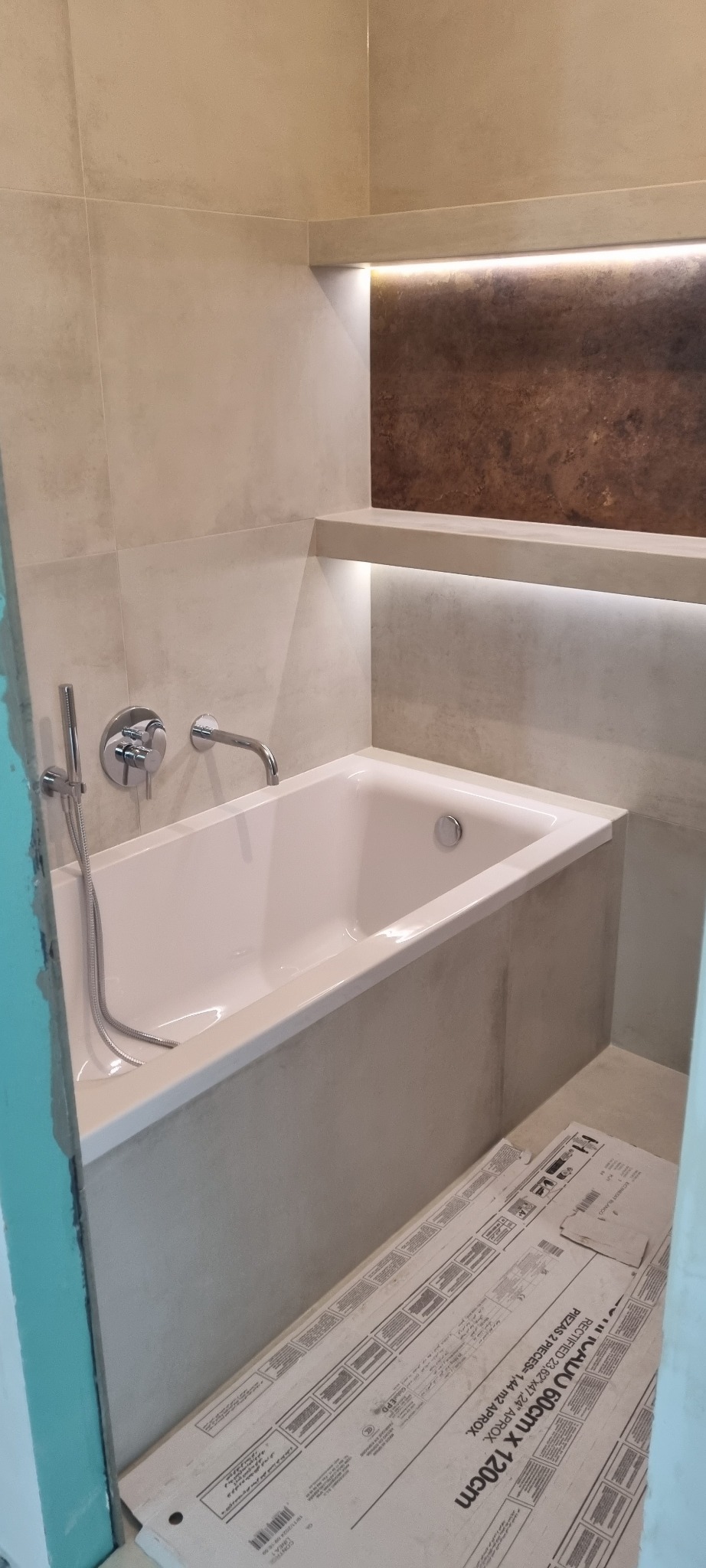 Nowoczesna łazienka w trakcie wykańczania: wanna obudowana płytkami, widoczna armatura, półka z oświetleniem LED, na podłodze opakowania płytek 60x120 cm.