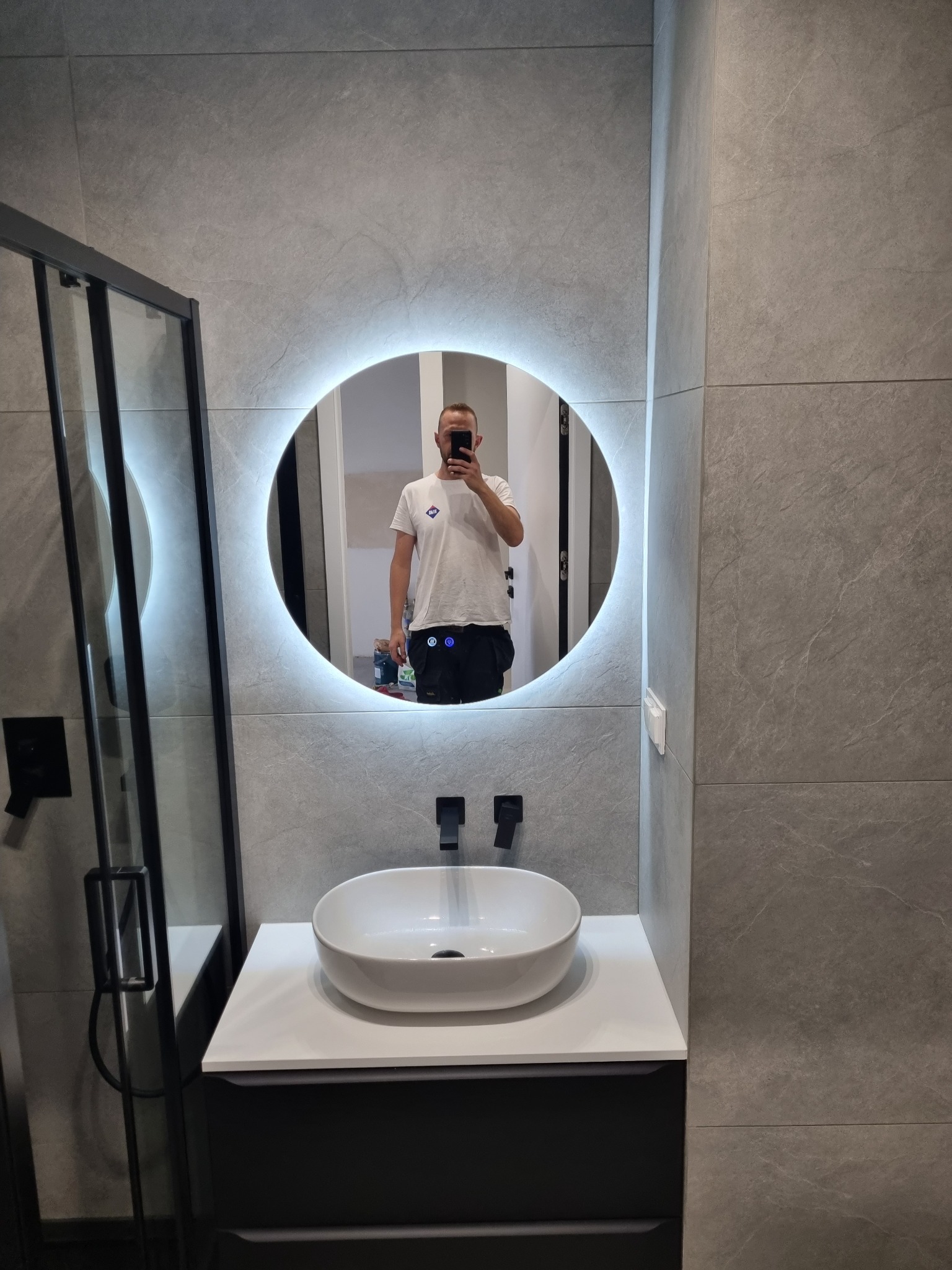 Nowoczesna łazienka z szarą płytką imitującą kamień, okrągłe lustro z podświetleniem LED i białą umywalką nablatową z czarną armaturą. Kabina prysznicowa z czarnymi profilami.