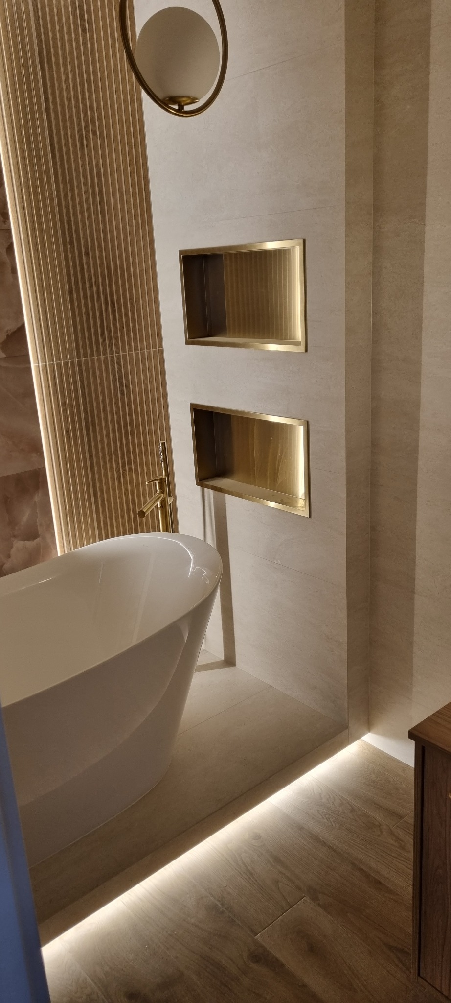 Nowoczesna łazienka: wolnostojąca wanna, złota armatura, wnęki ścienne z podświetleniem LED i drewniane panele ścienne. Subtelne oświetlenie podłogowe.
