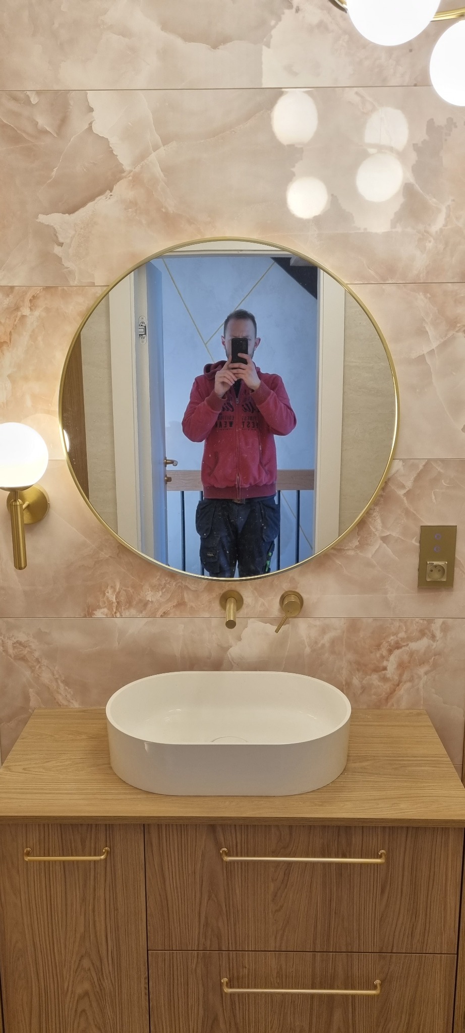 Ukończona łazienka z umywalką nablatową, złotą armaturą i lustrem. Widoczne płytki imitujące marmur i pracownik robiący zdjęcie w lustrze.