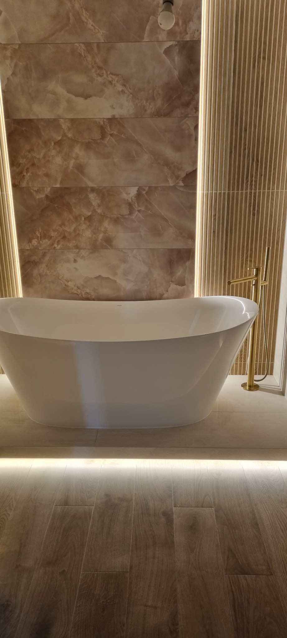 Nowoczesna łazienka z wolnostojącą wanną i podświetleniem LED, marmurowe płytki na ścianach i drewnopodobna podłoga. Złota bateria dodaje elegancji.
