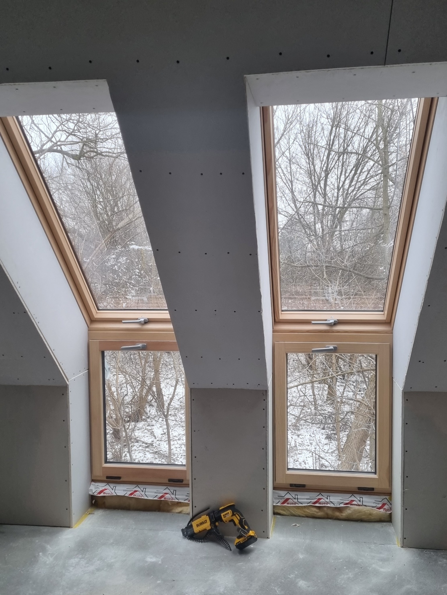 Poddasze z dwoma oknami dachowymi w trakcie montażu płyt kartonowo-gipsowych, widoczna wełna mineralna pod oknami i narzędzie do mocowania płyt.