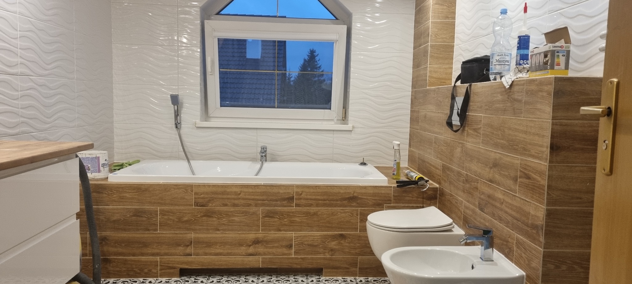 Ukończona łazienka z biało-brązowymi płytkami, białą wanną zabudowaną płytkami imitującymi drewno, toaletą, bidetem i oknem z widokiem na zewnątrz. Na wannie widoczne narzędzia i chemia budowlana.