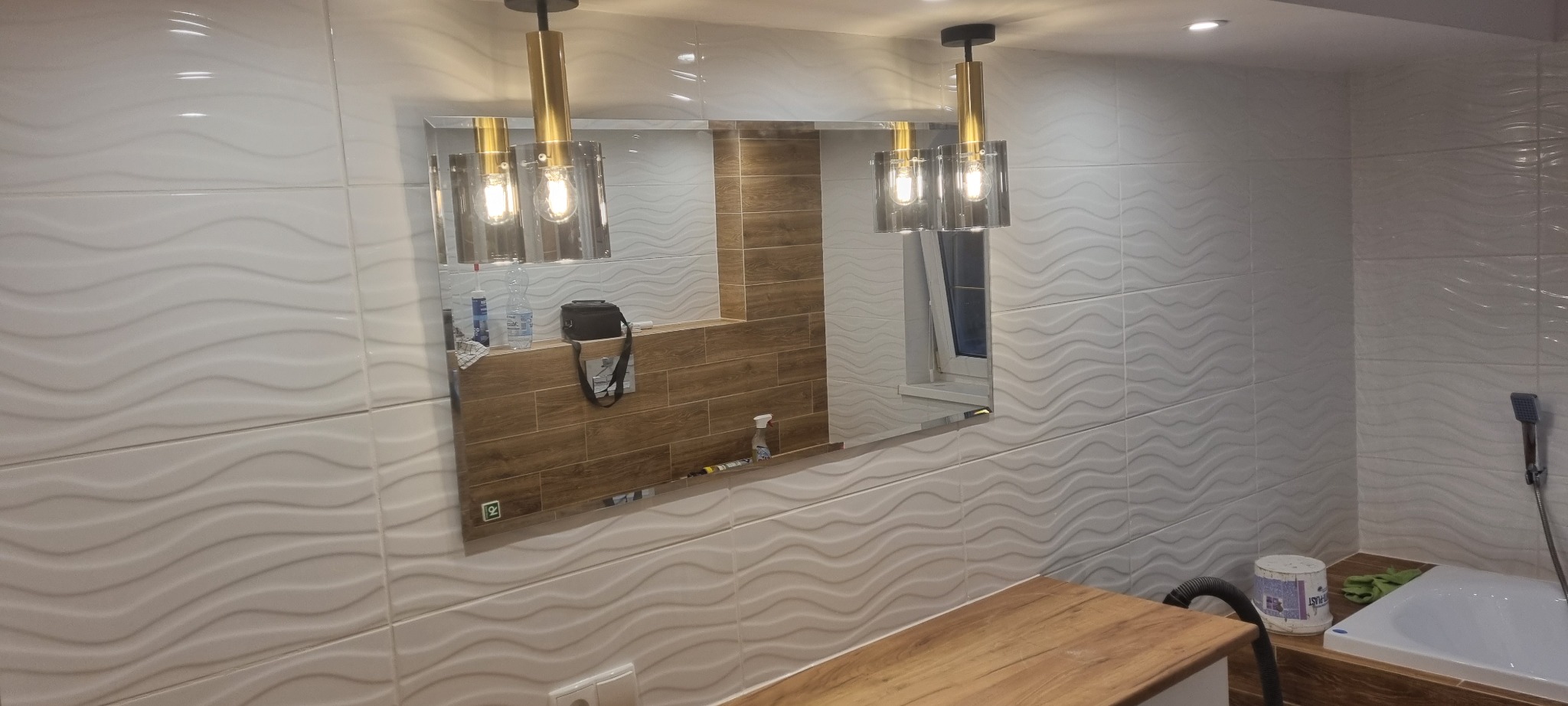 Nowoczesna łazienka z falistymi białymi płytkami, lustrem z odbiciem okna i drewnianej półki, oraz wiszącymi lampami z elementami złota.
