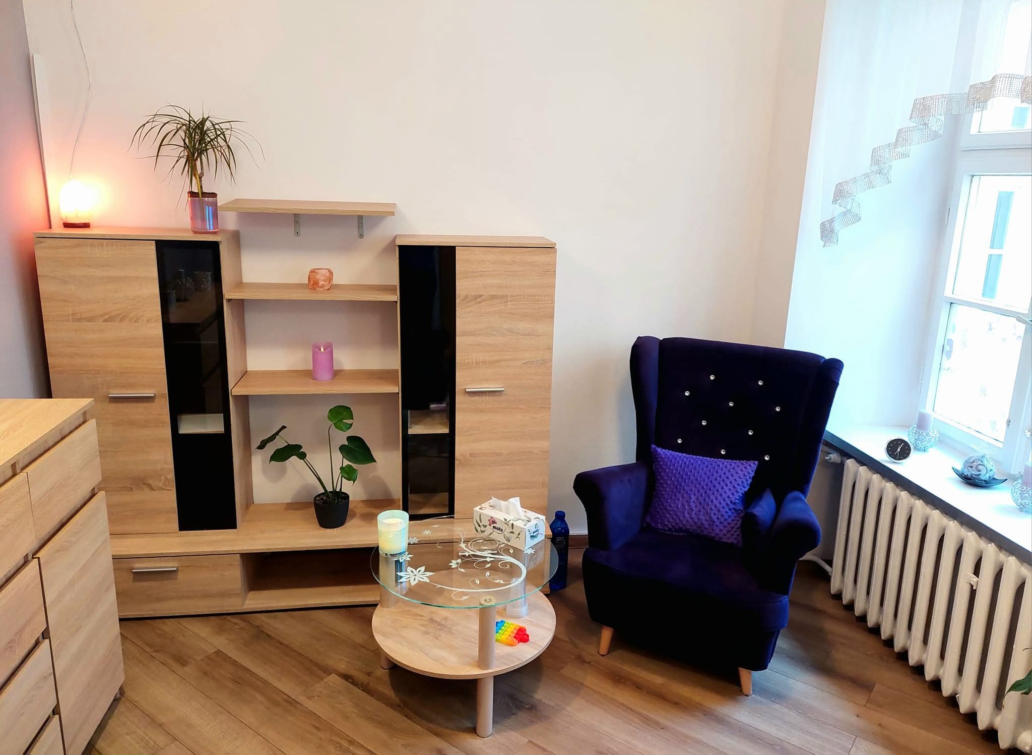 Jasne wnętrze gabinetu z drewnianymi meblami, fotelem w kolorze indygo i okrągłym stolikiem. Na parapecie dekoracje okienne i zegar.