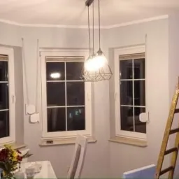 Świeżo pomalowane na szaro wnętrze pokoju z trzema oknami, drewnianą drabiną opartą o ścianę i wiszącą lampą z metalowym kloszem, dekorowane kwiatami na stole.