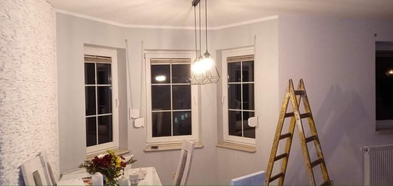 Świeżo pomalowane na szaro wnętrze pokoju z trzema oknami, drewnianą drabiną opartą o ścianę i wiszącą lampą z metalowym kloszem, dekorowane kwiatami na stole.