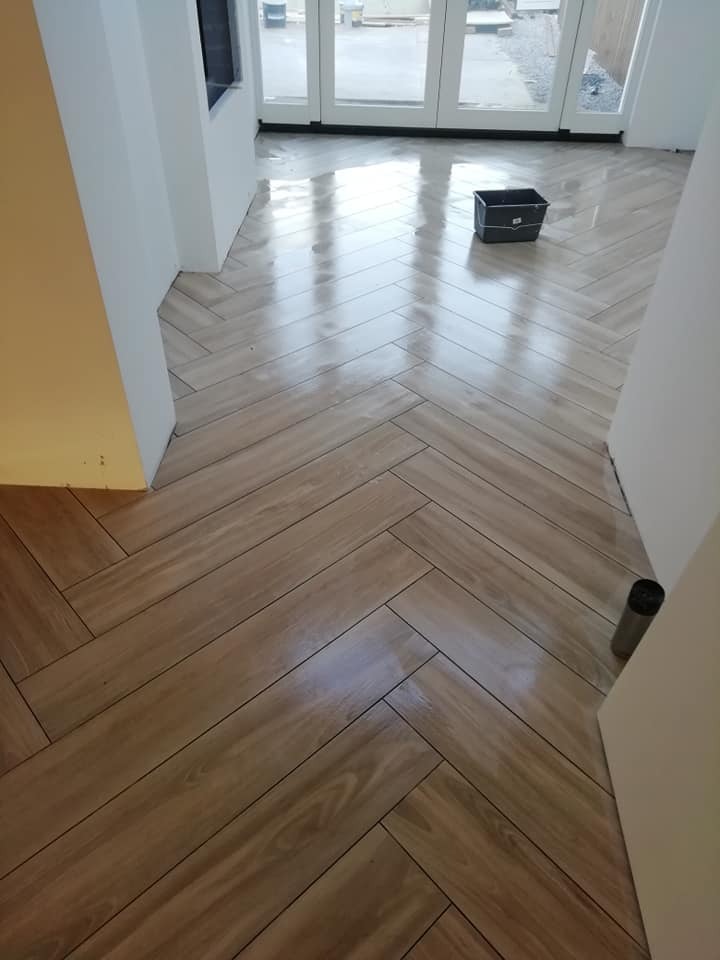 Ułożona pod kątem jodełki podłoga z paneli imitujących drewno w jasnym pomieszczeniu z widocznym przez szklane drzwi terenem budowy.