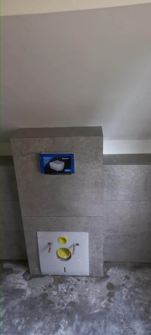 Szara ściana w łazience w trakcie montażu stelaża podtynkowego WC, widoczna instalacja z żółtymi elementami i pudełko z wizualizacją miski ustępowej.