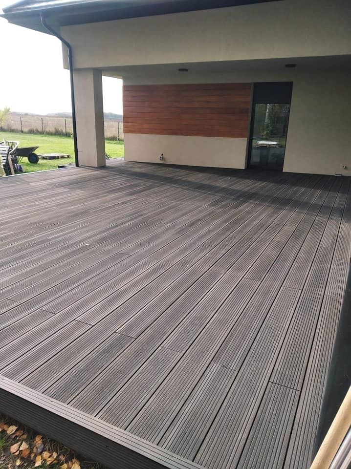 Szary taras z desek kompozytowych, z widokiem na trawnik i nowoczesny dom z drewnianą elewacją, w tle taczka z narzędziami.