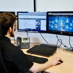 Mężczyzna siedzący przed dwoma monitorami, na których wyświetlane są strony internetowe, w tym jedna z grafiką sieci kontaktów, obok laptop, klawiatura, mysz i telefon stacjonarny na biurku.