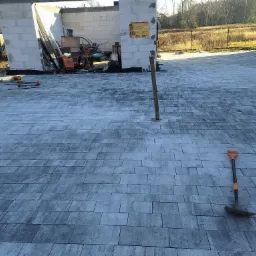 Ułożona kostka brukowa w szarych odcieniach na placu budowy z widoczną łopatą i paletami, w tle surowy budynek w trakcie wznoszenia.