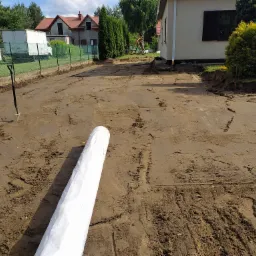 Przygotowany teren pod budowę chodnika z widocznymi śladami maszyn, rolka białej geowłókniny w pierwszym planie, w tle dom jednorodzinny i ogrodzenie.