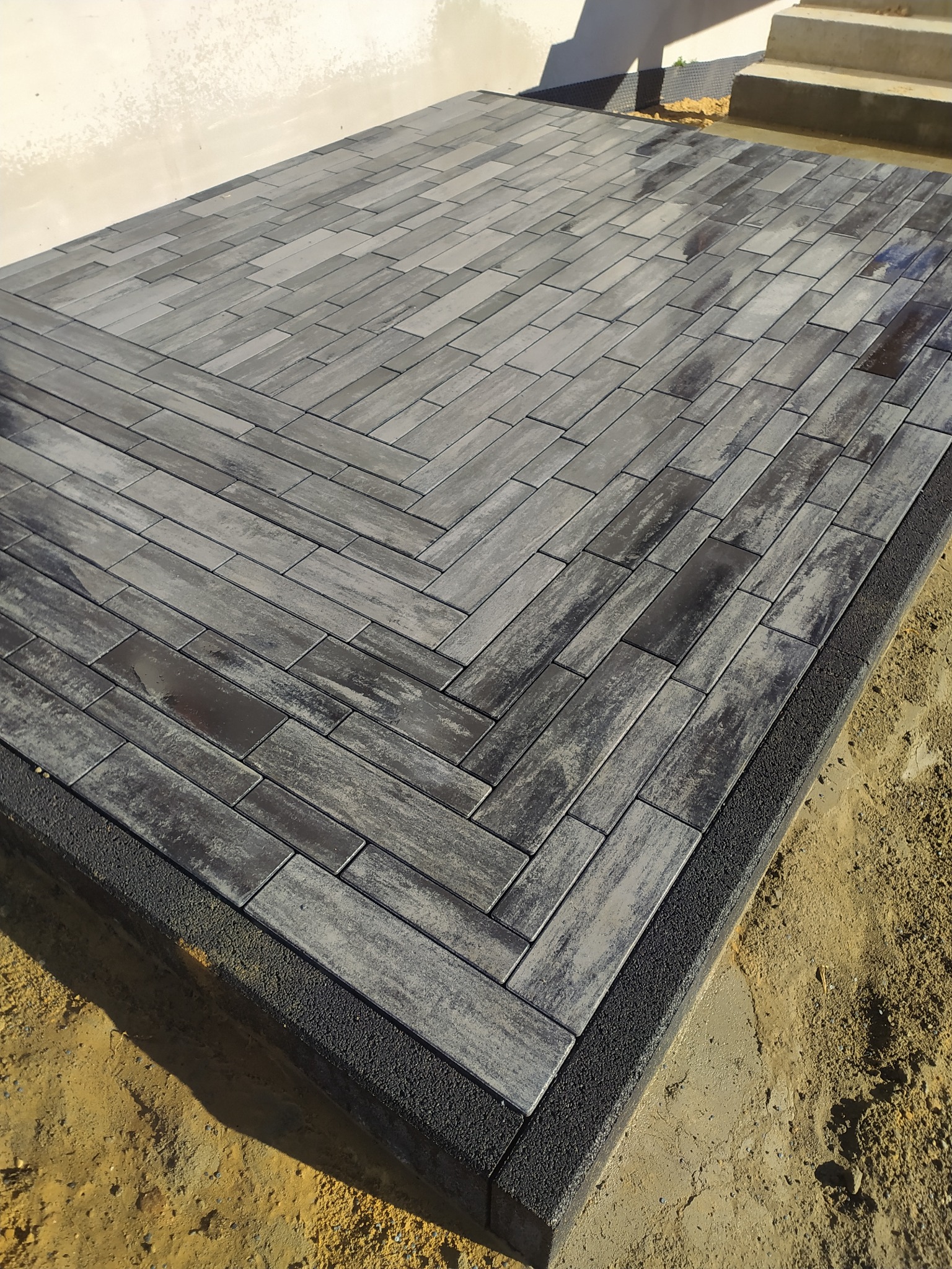 Ułożona, szara kostka brukowa w jodełkę na posesji w Kolbuszowej, obrzeża z ciemnej kostki, widoczny fragment budynku i schody.